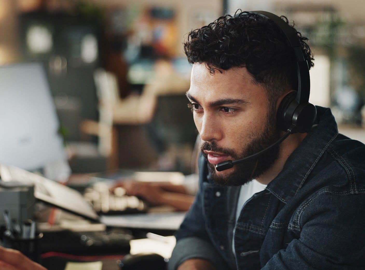Man with curly hair wearing a headset, focused on a computer screen in an office setting.