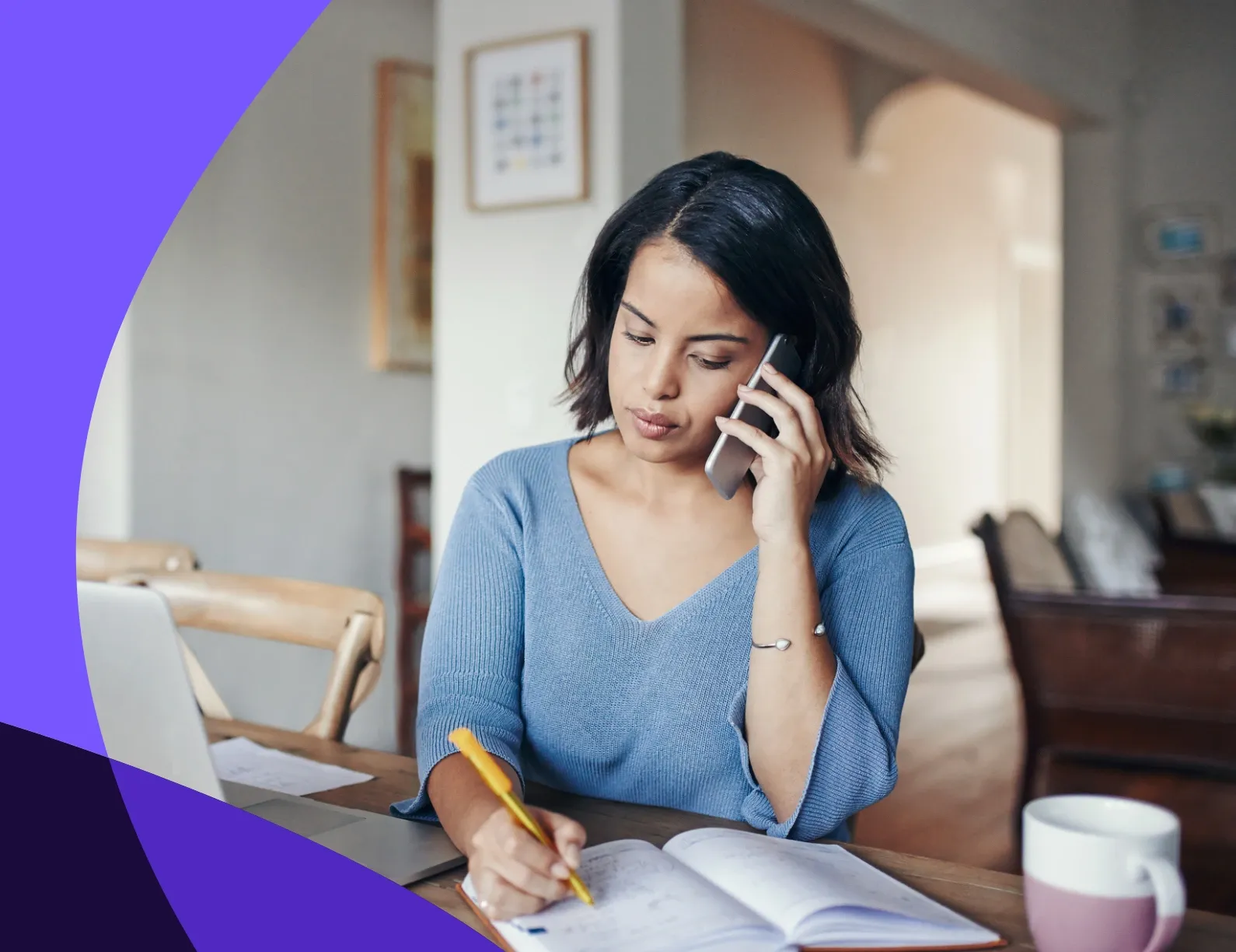 Woman in a blue sweater talking on the phone, taking notes at a table with a laptop and mug, in a well-lit room.