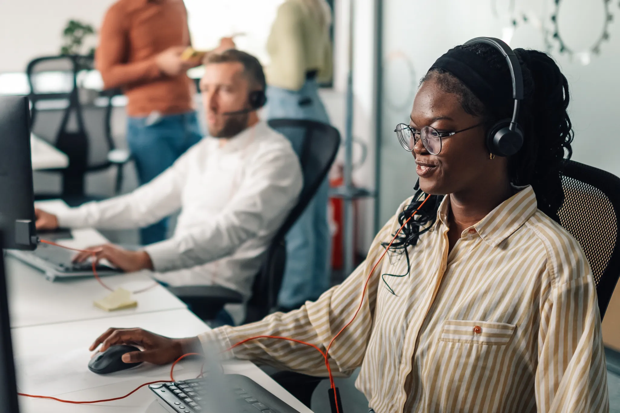 A woman wearing a headset works at a computer in an office, with a man in the background also on a headset.
