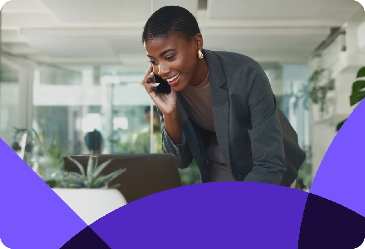 Smiling woman in a gray blazer talks on a phone while looking at a laptop in a modern office setting with plants.