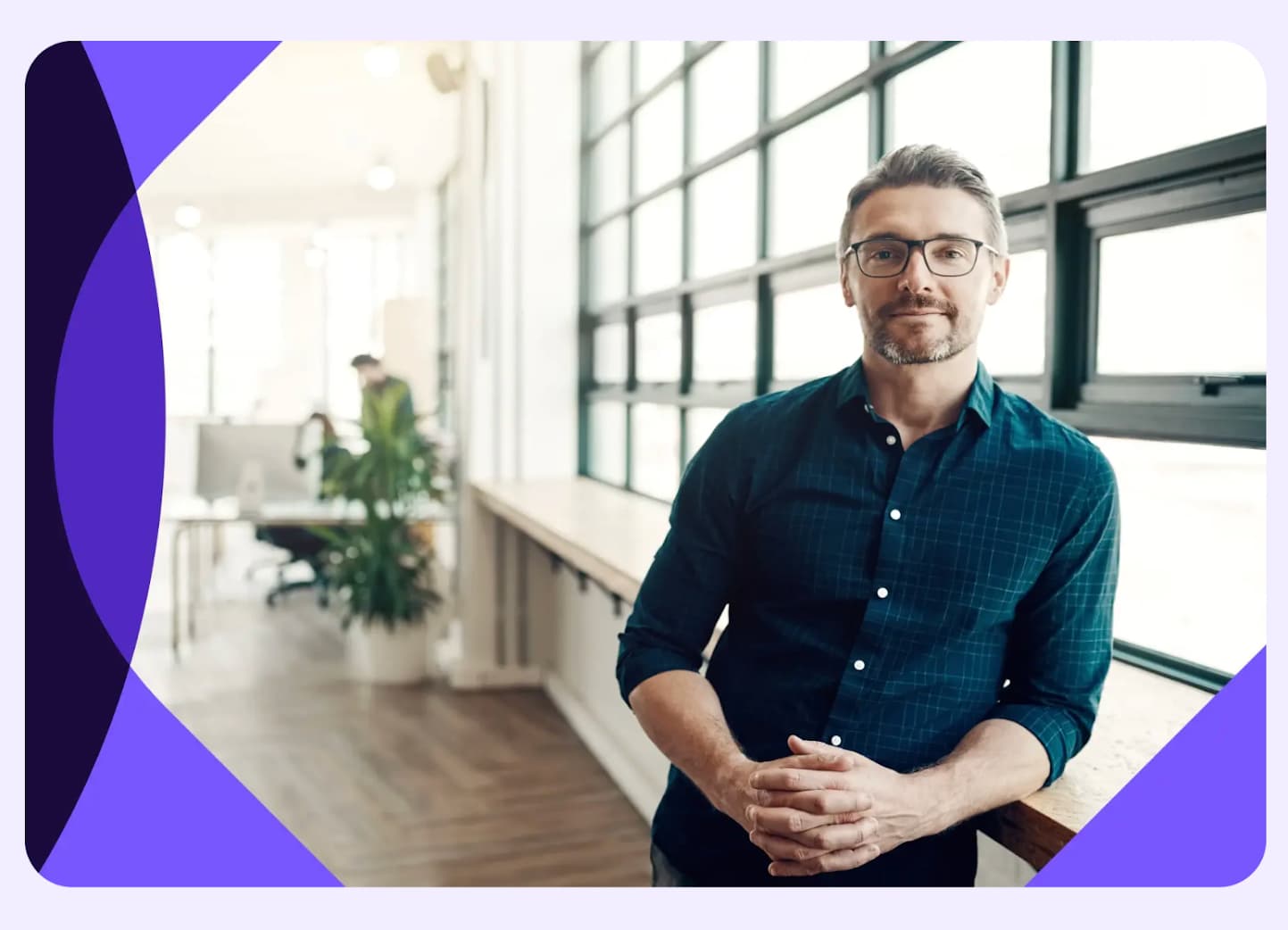 Man with glasses and a beard stands in a modern office, wearing a dark shirt. Large windows and a plant are in the background.