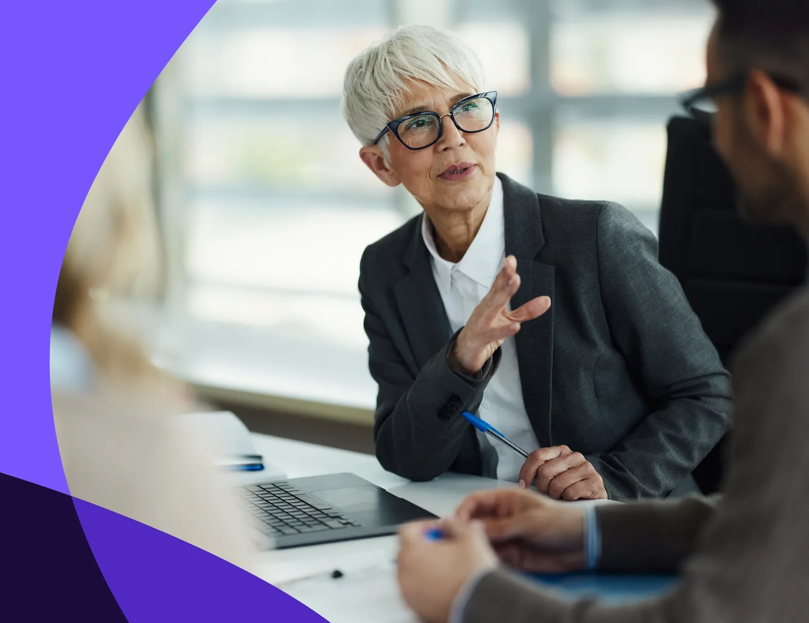 Elderly woman in a suit, holding a pen, discusses with colleagues at a table, with laptops and documents, in a bright office setting.