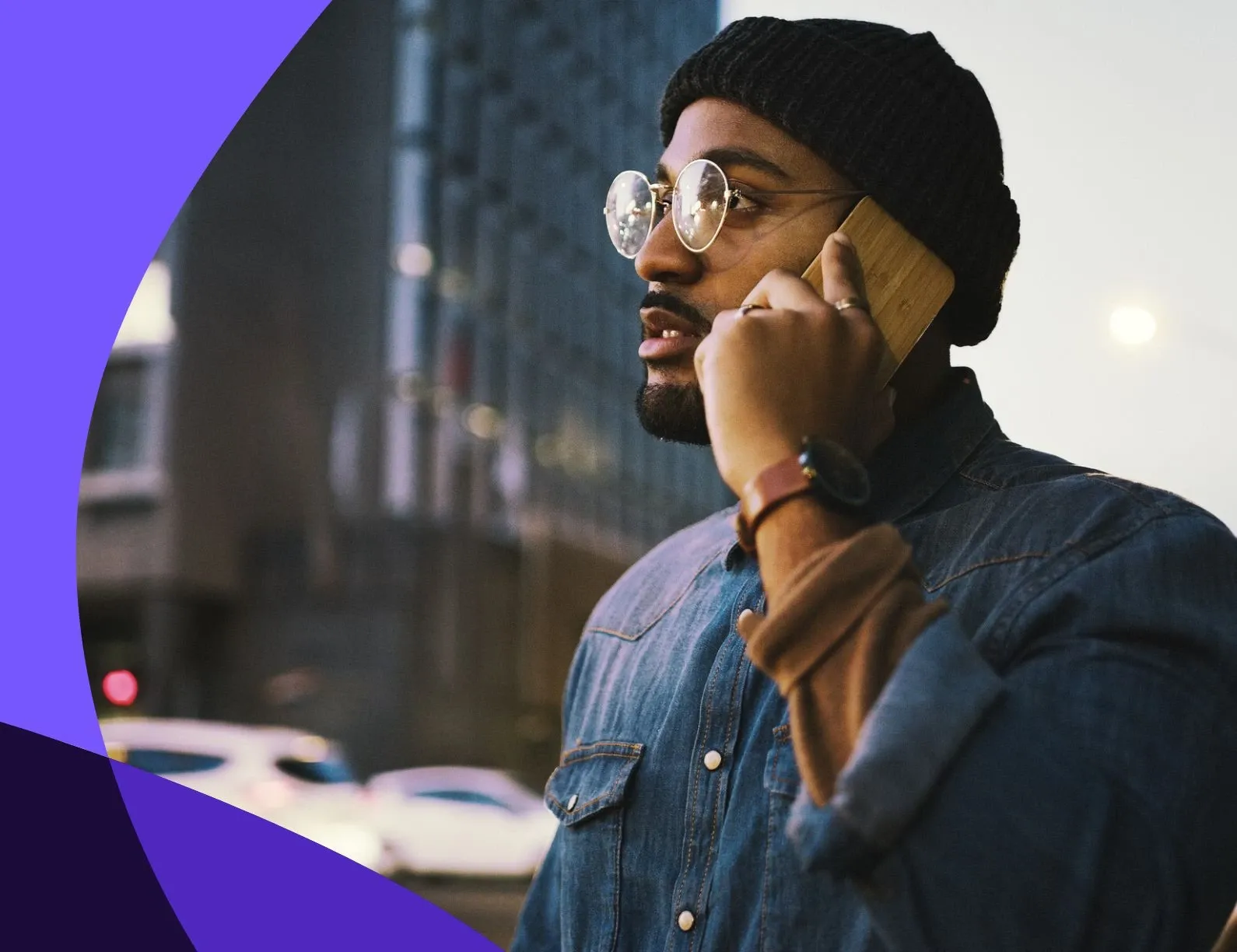 Man in denim jacket and beanie talks on phone on city street, wearing glasses and watch, with blurred buildings and cars in background.