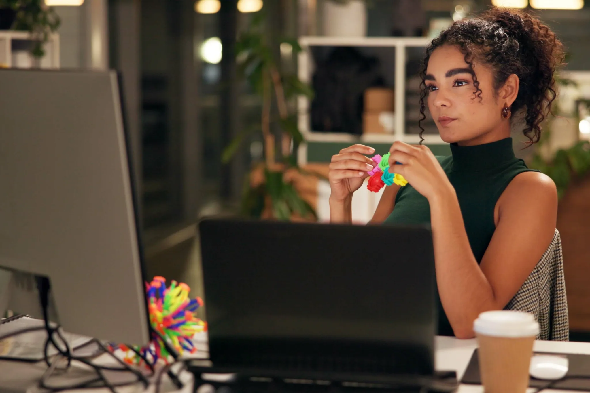 Woman with curly hair sits at desk, holding a colorful stress ball, looking at a computer screen. A coffee cup is beside her.
