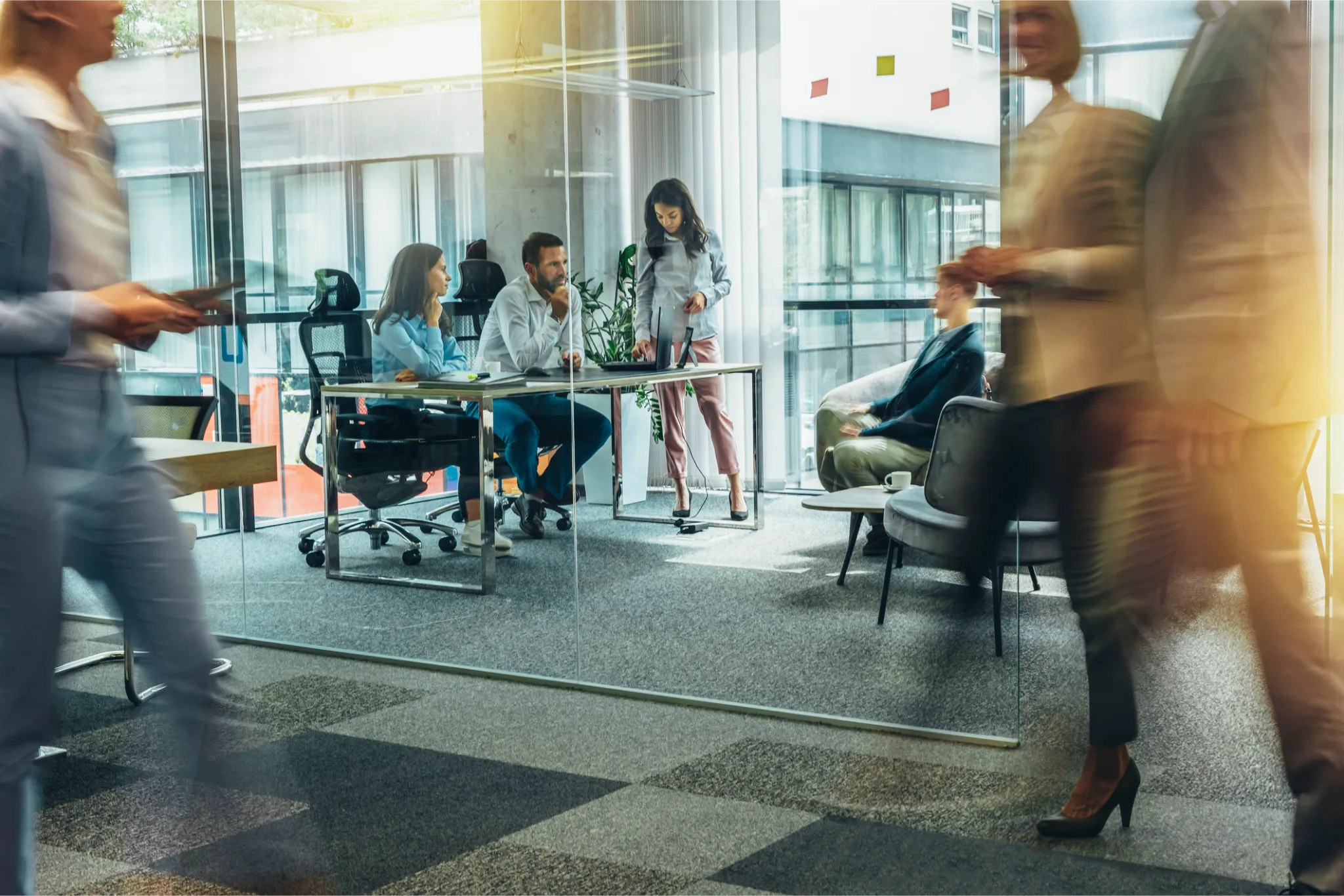 Modern office with glass walls; people working at desks and walking, creating a dynamic, professional atmosphere with natural light.