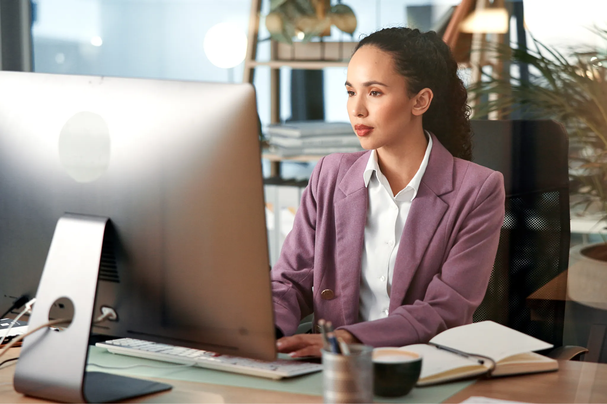 Woman in a purple blazer working at a computer in an office, surrounded by papers and plants.