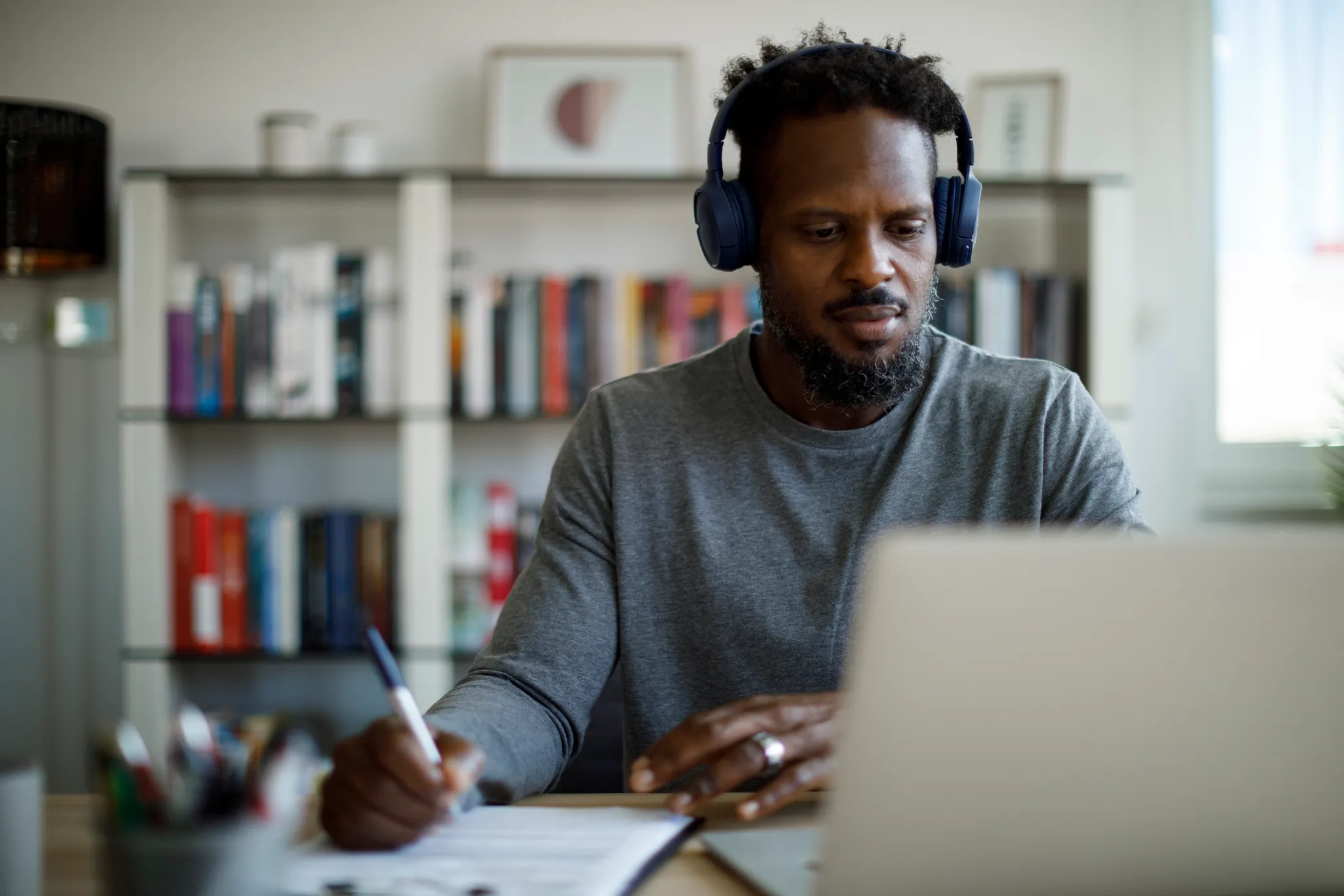 Man wearing headphones, writing on paper while using a laptop at a desk. Bookshelves and framed art are in the background.